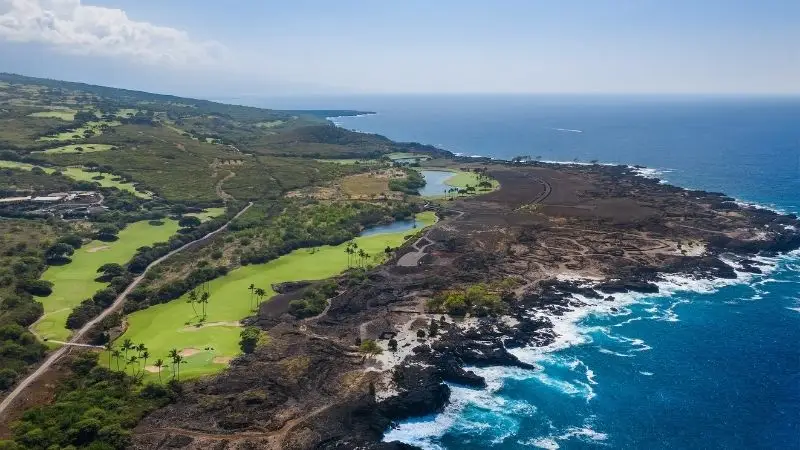 Aerial view of a coastal landscape featuring lush green golf courses, rocky volcanic terrain, and the blue ocean on the right. The shoreline has rugged rocks and waves crashing against them. The inland area is dotted with sparse vegetation and a few buildings.