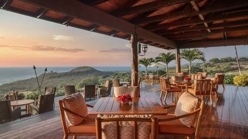 A scenic outdoor dining area features wooden tables and wicker armchairs under a rustic wooden roof. The area overlooks a stunning coastal landscape with lush greenery, hills, and the ocean in the background during sunset.