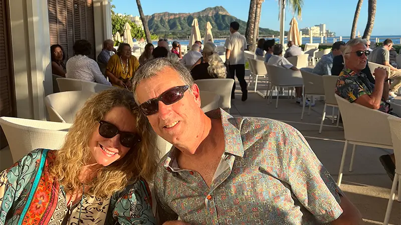 A smiling couple wearing sunglasses sit close together at an outdoor restaurant with people around them, epitomizing the joy of Why Waikiki. Palm trees, an ocean view, and a mountain in the background suggest a stunning tropical location.