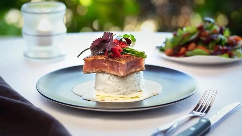 A gourmet dish featuring a perfectly seared piece of fish sits on a bed of creamy sauce atop a rice tower, garnished with microgreens, thinly sliced red radishes, and a single green pepper. This elegant creation is reminiscent of the best Hawaiian food Kauai offers, with an exquisite table setting including utensils and a side salad in the background.