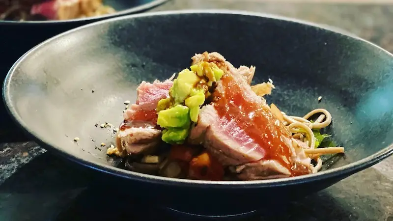 A close-up of a black bowl containing a gourmet dish, celebrated as some of the best Hawaiian food Kauai offers. The meal features seared slices of tuna topped with avocado chunks and garnished with a drizzle of sauce. Thin noodles and vegetables can be seen underneath the fish, arranged artfully.