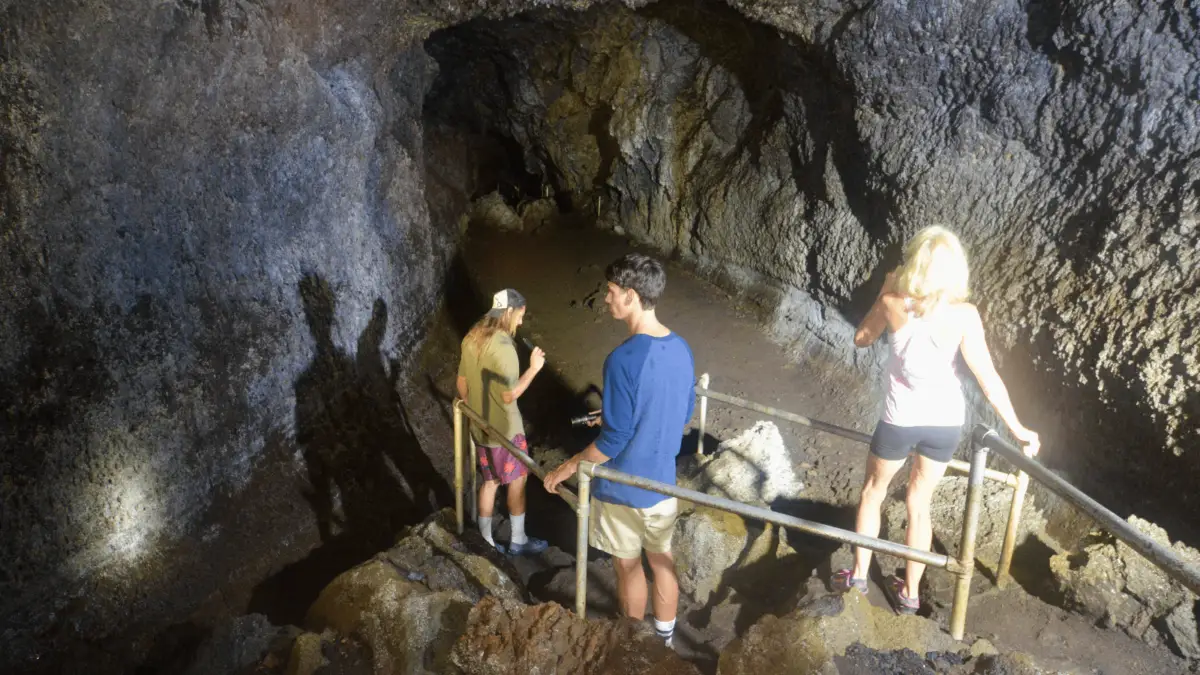 Three people descend a staircase into a dark, rocky cave in the Kaeleku Caverns of Hana. The cave's walls and floor are rough and uneven. The person on the left wears a hat and shorts, the person in the middle wears a blue shirt and shorts, and the person on the right sports a white tank top and shorts.