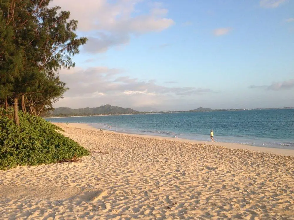 A serene view of a sandy beach with clear blue waters stretching into the horizon. A few people are walking along the shoreline, enjoying one of the best beaches for long walks on Oahu. Trees and greenery border the beach on the left side, and distant hills are visible under a partly cloudy sky.