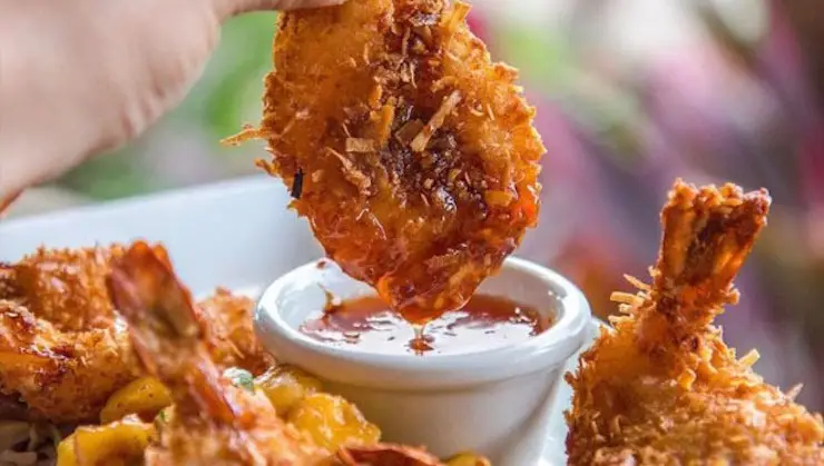 A hand holding a piece of crispy coconut shrimp, dipping it into a small cup of red sauce. Other pieces of coconut shrimp are visible on the plate, creating a mouthwatering and appetizing presentation. The background is blurred, keeping the focus on one of the best Hawaiian foods Kauai has to offer.