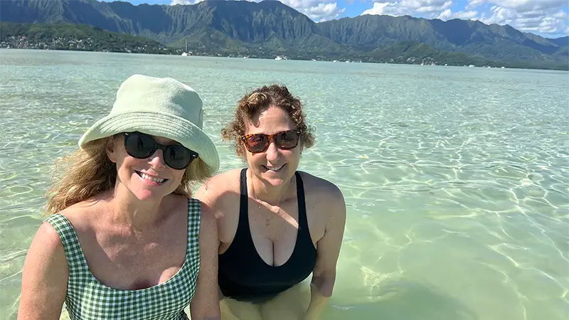 Two women, one in a checkered swimsuit and sun hat, and the other in a black swimsuit, are standing in shallow clear water with mountains and a bright sky in the background. Both are smiling and wearing sunglasses. This scene perfectly captures why Waikiki is a favorite vacation spot.