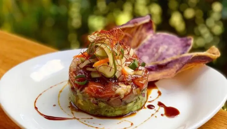 A circular mound of poke, layered with diced fish, avocado, and vegetables, drizzled with a dark sauce, garnished with shredded carrots and herbs, served on a white plate with two large purple yam chips in the background. This dish exemplifies the best Hawaiian food Kauai has to offer in an inviting outdoor setting.