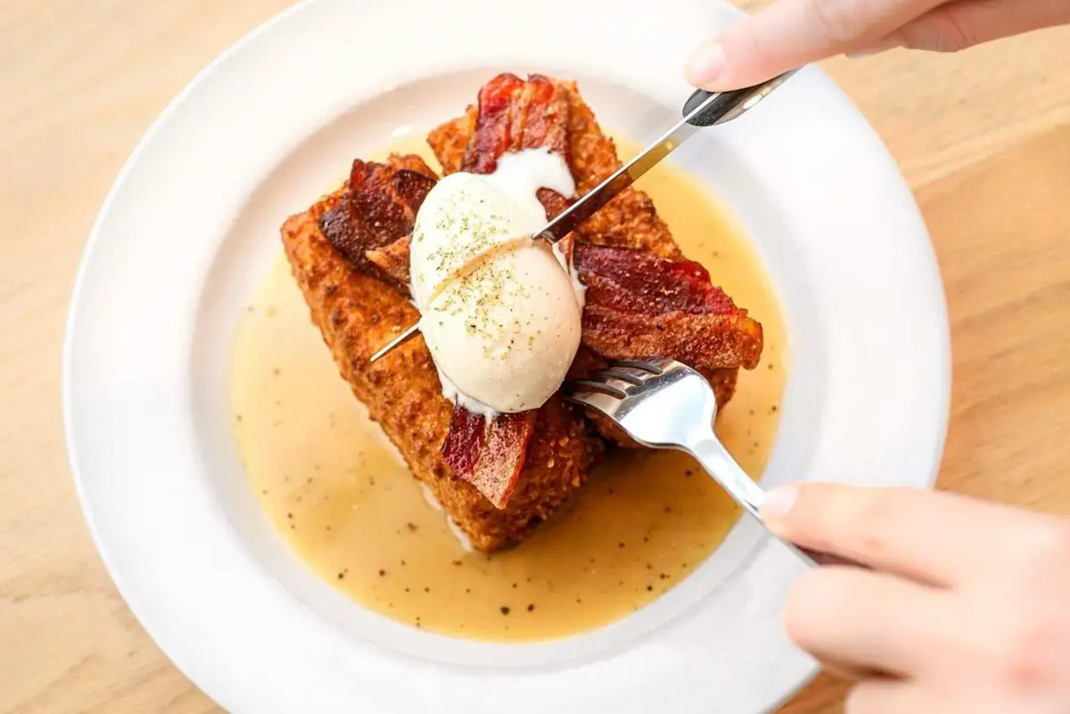 A person is cutting into a dish with a knife and fork. The dish, crafted by Maui Chef Lee Anne Wong, features French toast topped with crispy bacon and a poached egg, drizzled with syrup, and sprinkled with pepper, served on a white plate. The background is a wooden surface.