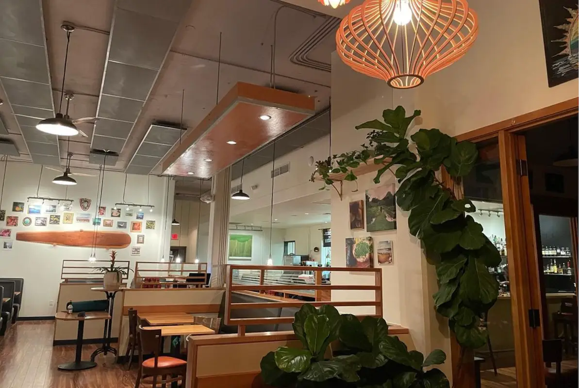 A cozy restaurant interior with wooden tables and booths, green plants, and warm hanging lights. The walls are adorned with various framed pictures and paintings. A surfboard decoration is visible on one wall, showcasing a nod to Maui Chef Lee Anne Wong’s coastal influences. The bar area is partially visible in the background.