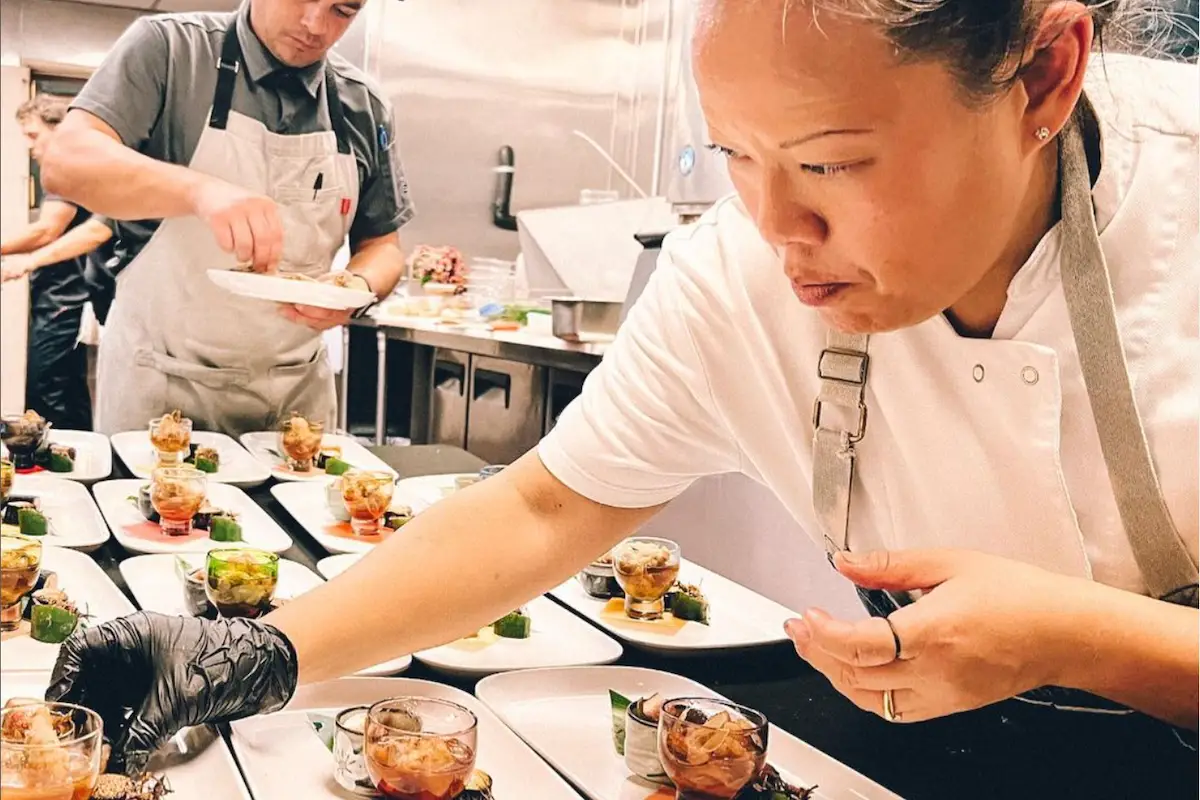 Maui Chef Lee Anne Wong, in a white uniform, carefully plates food in a busy kitchen while a male chef in the background, also in an apron, prepares another dish. Multiple small glass bowls with food are arranged on the counter in front of them.