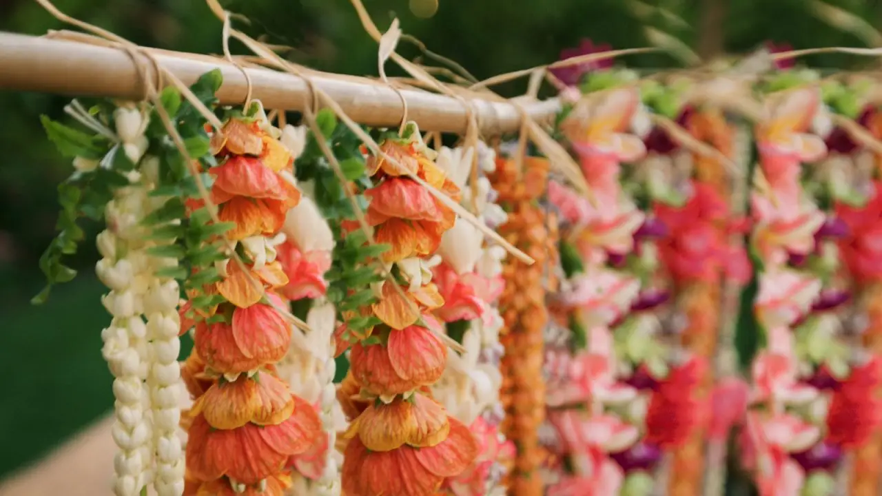 A close-up of multiple colorful flower garlands hanging from a rod, perfect inspiration for those looking to make a lei. The garlands feature a combination of orange, white, red, purple, and green flowers, creating a vibrant and festive display against a blurred background.