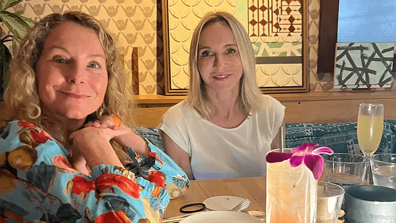 Two women seated at a restaurant table in Waikiki smile at the camera. They are positioned side by side, with one in a colorful top and the other in a plain white shirt. On the table in front of them are drinks, including a cocktail adorned with a purple orchid.