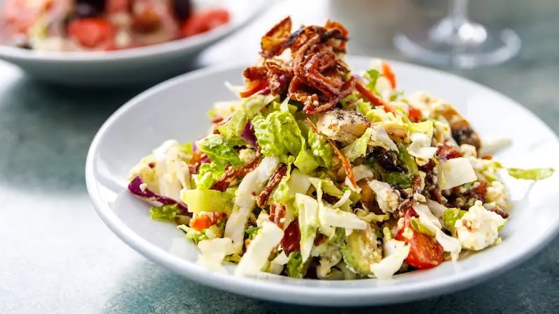 A close-up of a fresh, colorful salad on a white plate at the best Gluten-Free Wailea spot. The salad includes a mixture of greens, diced vegetables, and crispy bacon bits on top. The background features another plate with additional food items blurred out. The table has a light greenish hue.
