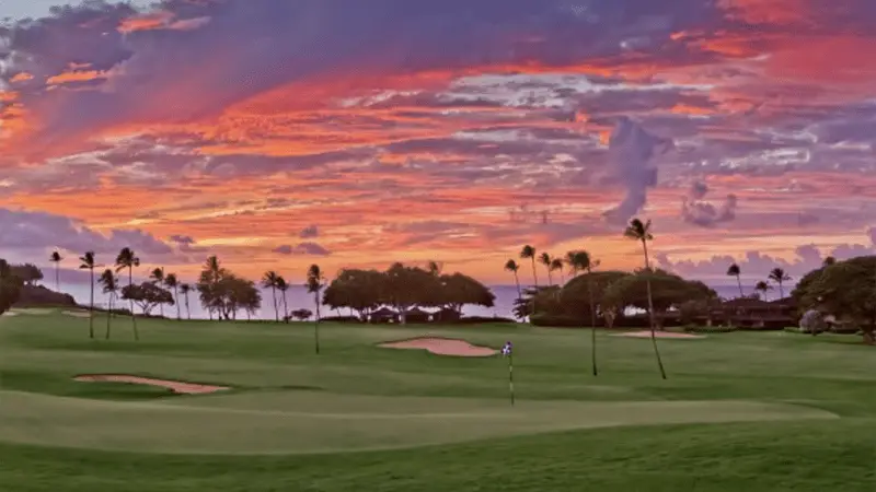 Vibrant sunset sky with shades of orange, pink, and purple over one of the best golf courses in Maui. Palm trees and sand bunkers are scattered across the lush, green course while the silhouette of trees is visible in the background near calm ocean waters.