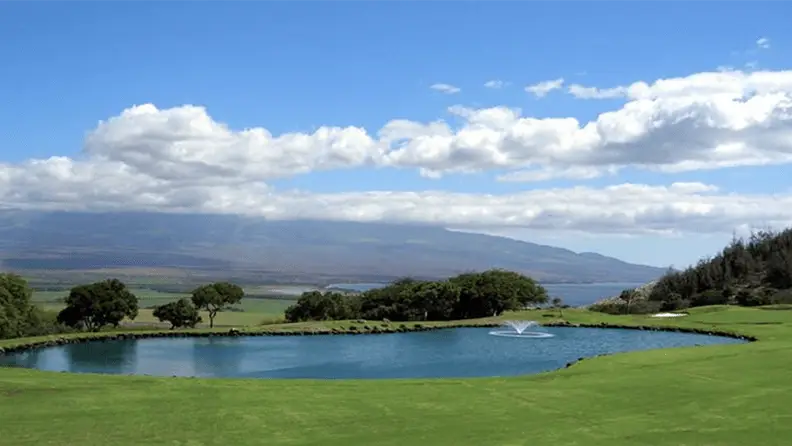 A serene golf course, often hailed as one of the best golf courses in Maui, features a clear pond with a small fountain in the center. The pond is surrounded by lush green grass, trees, and rolling hills. In the background, mountains stretch under a blue sky with scattered clouds.
