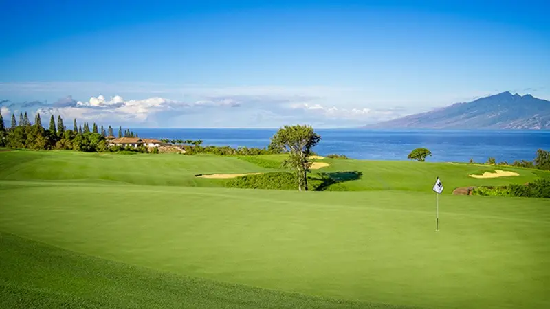 A picturesque golf course featuring a well-maintained green, a flag in the hole, and sand bunkers in the distance. Often hailed as one of the best golf courses on Maui, it overlooks a serene blue ocean, with mountainous terrain and a partly cloudy sky in the background.