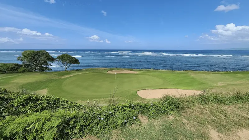 A picturesque coastal golf course, touted as one of the best golf courses Maui has to offer, with a putting green and sand bunker in the foreground. The lush green grass contrasts with the clear blue ocean and sky in the background. Waves gently lap along the shore, and trees frame the left side of the scene.