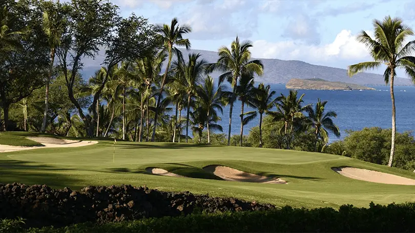 A scenic view of one of the best golf courses Maui has to offer, showcasing a lush green golf course with palm trees. Sand bunkers dot the landscape, while the ocean glistens in the background with an island visible in the distance. The sky is partly cloudy, adding to its serene beauty.