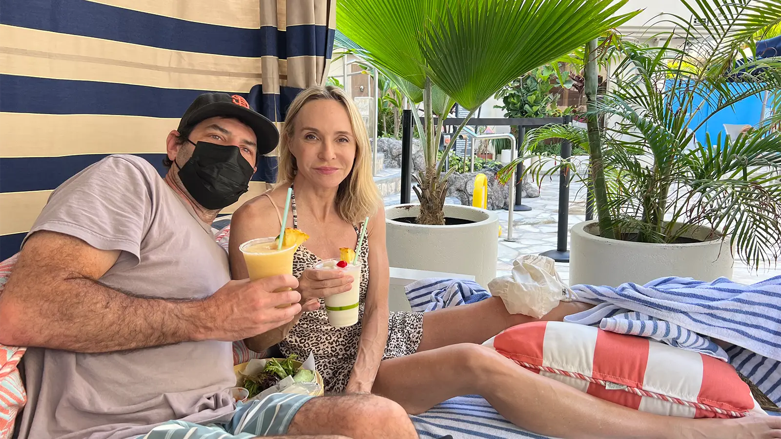 A man and a woman relax on lounge chairs under a cabana with striped curtains, surrounded by potted plants. The man wears a black cap, a face mask, and holds a blended drink, while the woman in a patterned dress enjoys her tropical drink. Towels and cushions are visible, embodying the essence of Why Waikiki.