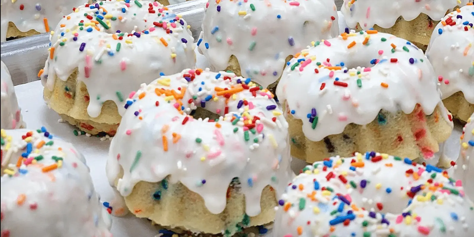 Small bundt cakes are decorated with white icing and colorful sprinkles. These best desserts in Honolulu are neatly arranged on a tray, showcasing their vibrant and festive appearance. The icing drips slightly over the sides, adding a playful touch.