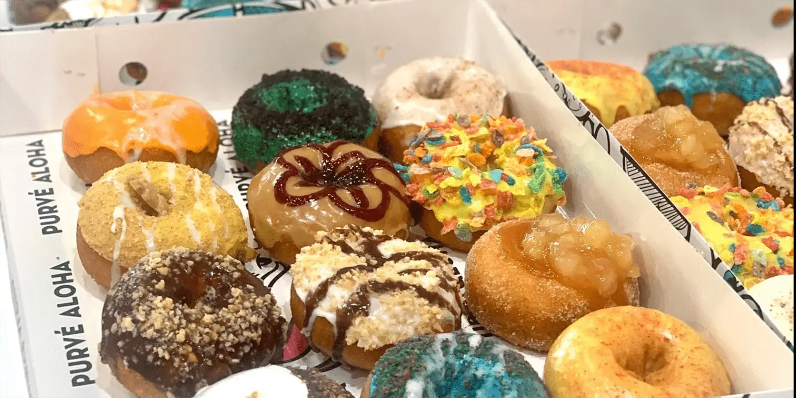 A box filled with assorted gourmet mini donuts, each topped with a variety of colorful and creative toppings like fruit loops, caramel drizzle, chocolate, blue and yellow sprinkles, and crushed nuts. The box liner reads "PURVÉ ALOHA." Discover the best desserts Honolulu has to offer.