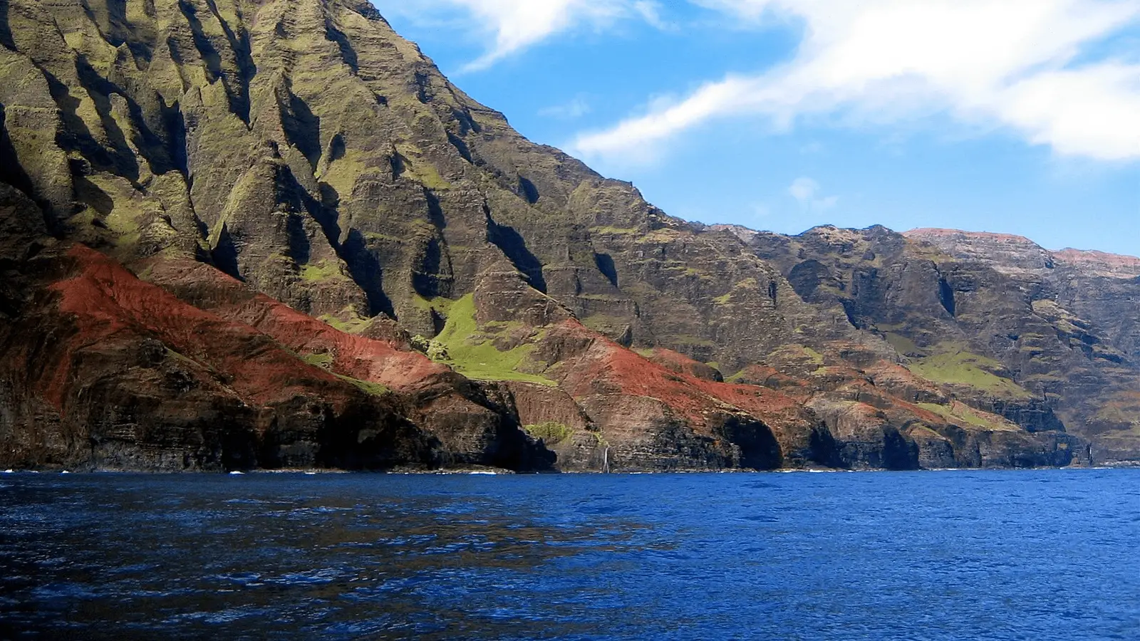 Image of a rugged coastal cliff, featuring layers of red, green, and brown rock formations. The bright blue ocean stretches out in front of the cliffs—one of the best places to scuba dive on Kauai. The sky above is partly cloudy with patches of blue peeking through, creating a striking contrast.