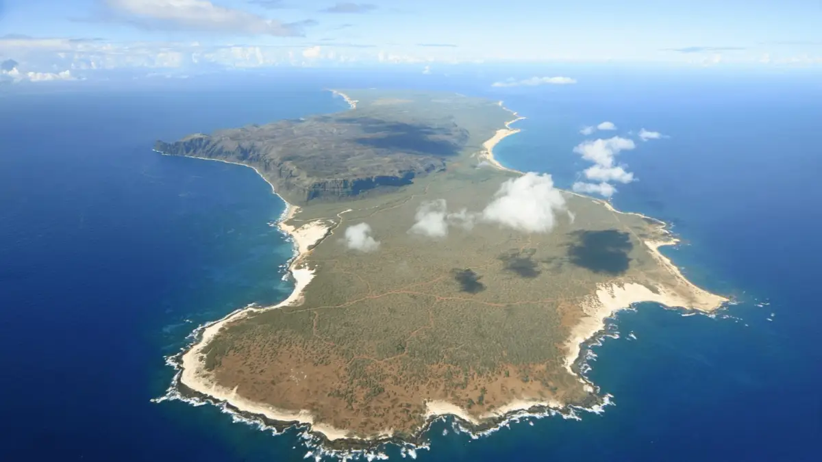 Aerial view of a vast, remote island surrounded by clear blue ocean waters. The island features rugged terrain with patches of greenery and brownish soil, while edges are lined with rocky cliffs and white sandy textures. A few clouds hover over the landscape—one of the best places to scuba dive on Kauai.