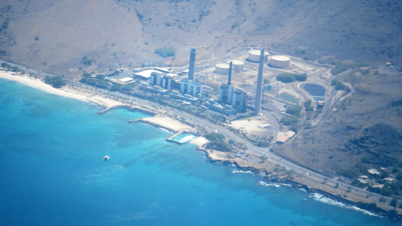 Aerial view of a coastal industrial facility with multiple buildings and tall chimneys located near a sandy beach and turquoise blue ocean. Hills and rocky terrain surround the area. A road runs along the coastline, providing easy access for those heading to scuba diving spots in Oahu. A small pool is visible near the beach.