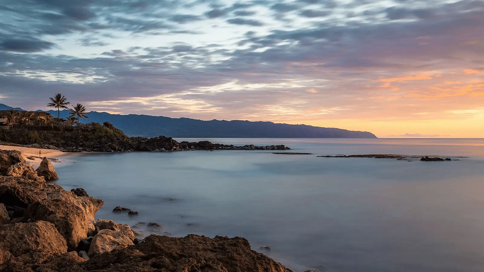 A tranquil beach scene at sunset, featuring a rocky shoreline on the left, calm waters, and a sky adorned with soft clouds illuminated by orange and pink hues. Palm trees and distant mountains enhance the serene landscape on the horizon, perfect for reflecting after an exhilarating day of scuba diving Oahu.