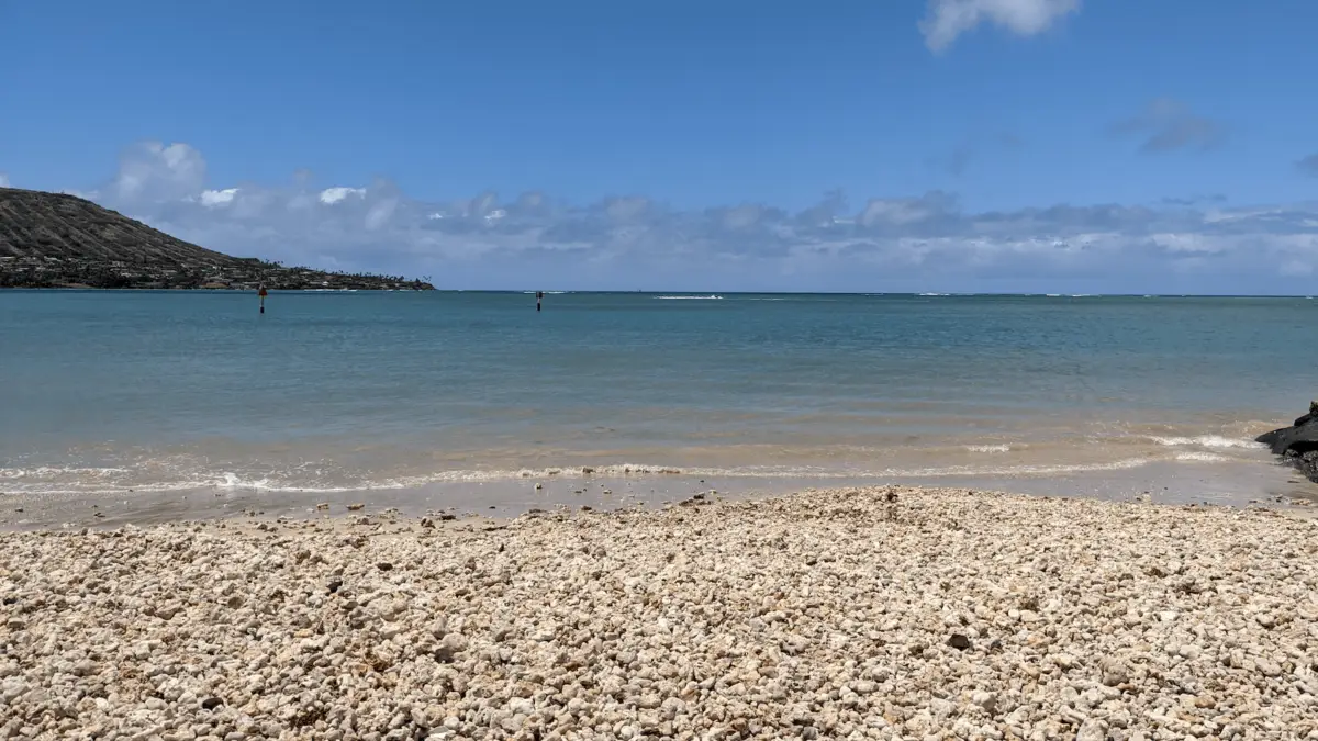 A rocky beach with white stones stretches out to meet calm, clear blue waters under a bright sky, perfect for scuba diving. A distant headland with some vegetation is visible on the left side of the horizon, reminiscent of Oahu. Patches of clouds dot the otherwise clear sky.
