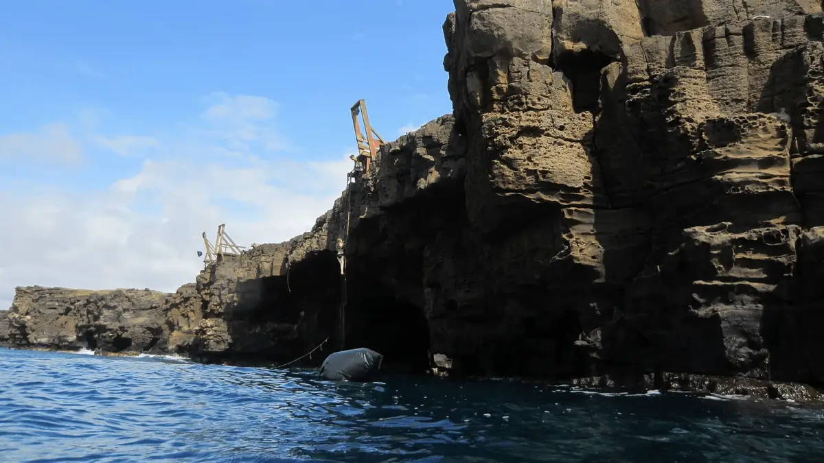 A rocky cliffside with distinct geological formations stands against a bright blue sky. A wooden ladder is attached to the cliff, leading to an enclosed area. Below, a dark rubber raft is tethered to the rocks, floating on the calm, deep blue water—one of the best places to scuba dive on the Big Island.