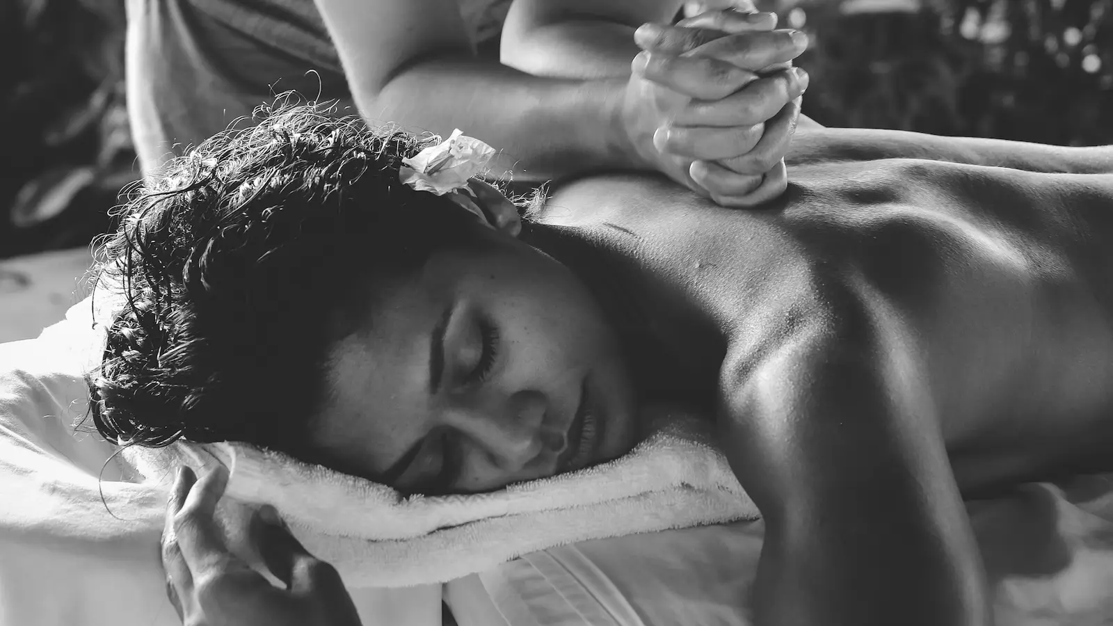 Black and white image of a person lying face down with eyes closed, receiving a back massage. Their head rests on a towel, and another person's hands are seen applying pressure to their back. The scene exudes relaxation and tranquility, highlighting the real health benefits of Hawaii’s ancient Lomilomi massage.