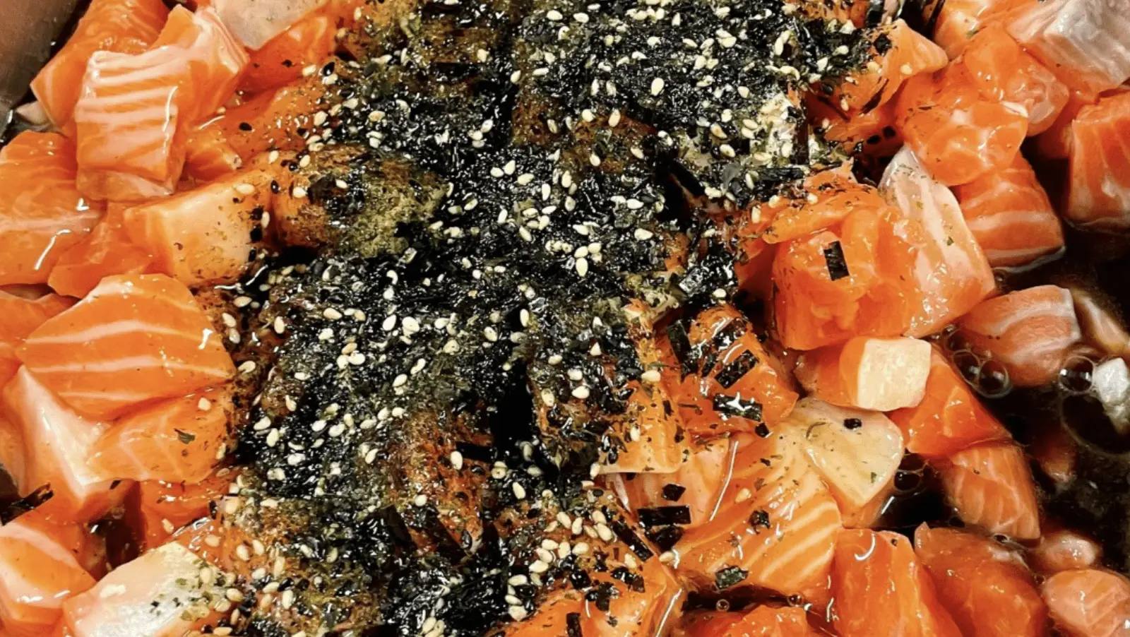 Close-up of a dish featuring diced raw salmon, sprinkled with shredded seaweed, sesame seeds, and spices. The vibrant orange color of the salmon contrasts with the dark green seaweed and white sesame seeds, creating a visually appealing presentation rooted in poke history.