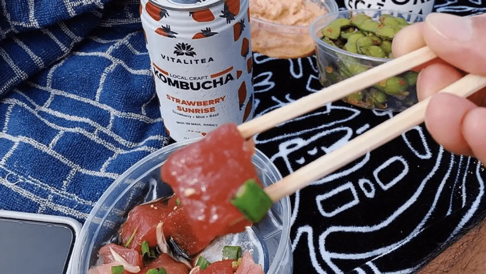 A close-up of chopsticks holding a piece of sashimi above a container of more sashimi. There's a can of strawberry kombucha, a towel with blue patterns, and containers of edamame and dipping sauce in the background, subtly hinting at poke history intertwined with modern flavors.