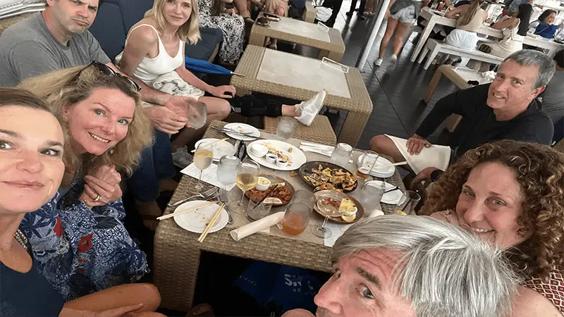 A group of six people sits around a table at a restaurant. The table is covered with empty plates, glasses, and leftover food. They are smiling and appear to be enjoying each other's company. In the background, more patrons and white interior furnishings give the scene a Why Waikiki vibe.