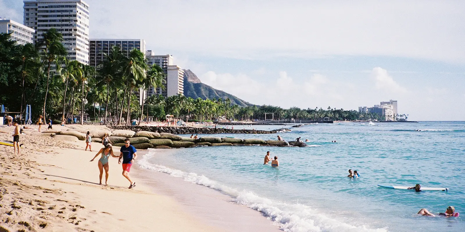 A family-friendly beach on Oahu, with people swimming and relaxing near the shore. A couple walks close to the waterline while others swim or paddle in the calm blue ocean. High-rise buildings and lush palm trees line the beach, with distant hills and a clear sky in the background.