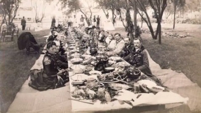 A historical black and white photo depicts a long outdoor banquet table laden with food and surrounded by people, reminiscent of a Hawaiian luau. Men and women dressed in formal attire are seated on either side of the table, which is set under trees in a park-like setting.