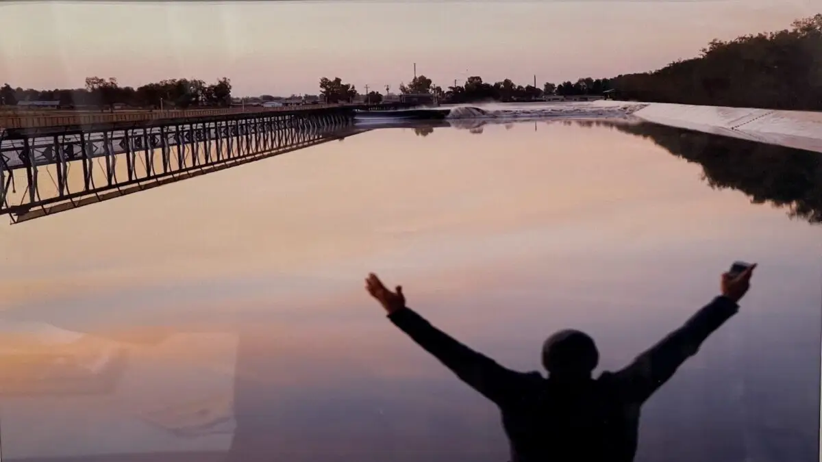 Person with outstretched arms stands on a bank overlooking a calm water surface at sunset. The sky is tinged with soft pinks and purples, reflected in the water—a perfect moment showcasing the benefits of life on the water. A bridge extends across the water, and distant trees line the horizon.