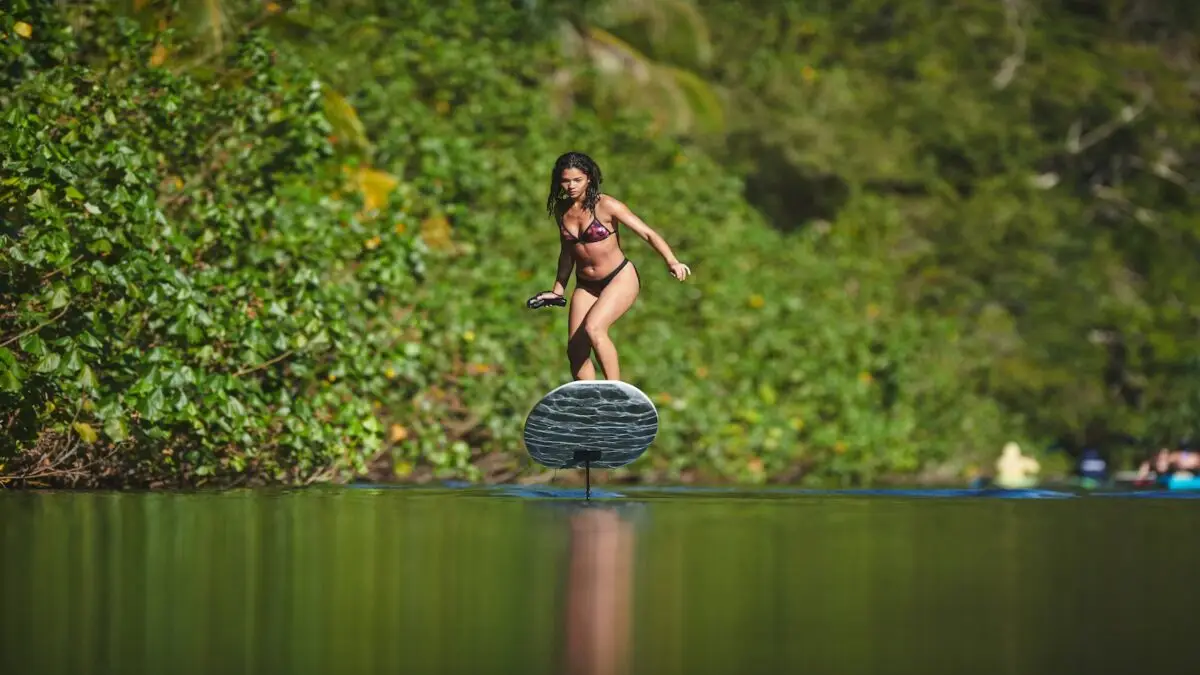 A woman in a red bikini rides a hydrofoil board on calm water surrounded by lush green vegetation. She maintains balance while holding something in her hand, embodying the benefits of life on the water. The background is filled with greenery, suggesting a tropical environment.