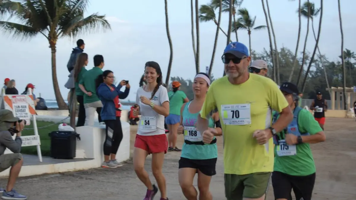 A group of runners, wearing race bibs and athletic clothing, participate in a North Shore annual event near the coast. Palms and spectators are visible in the background. One runner wears a neon yellow shirt and blue cap, while others are in various casual sportswear.