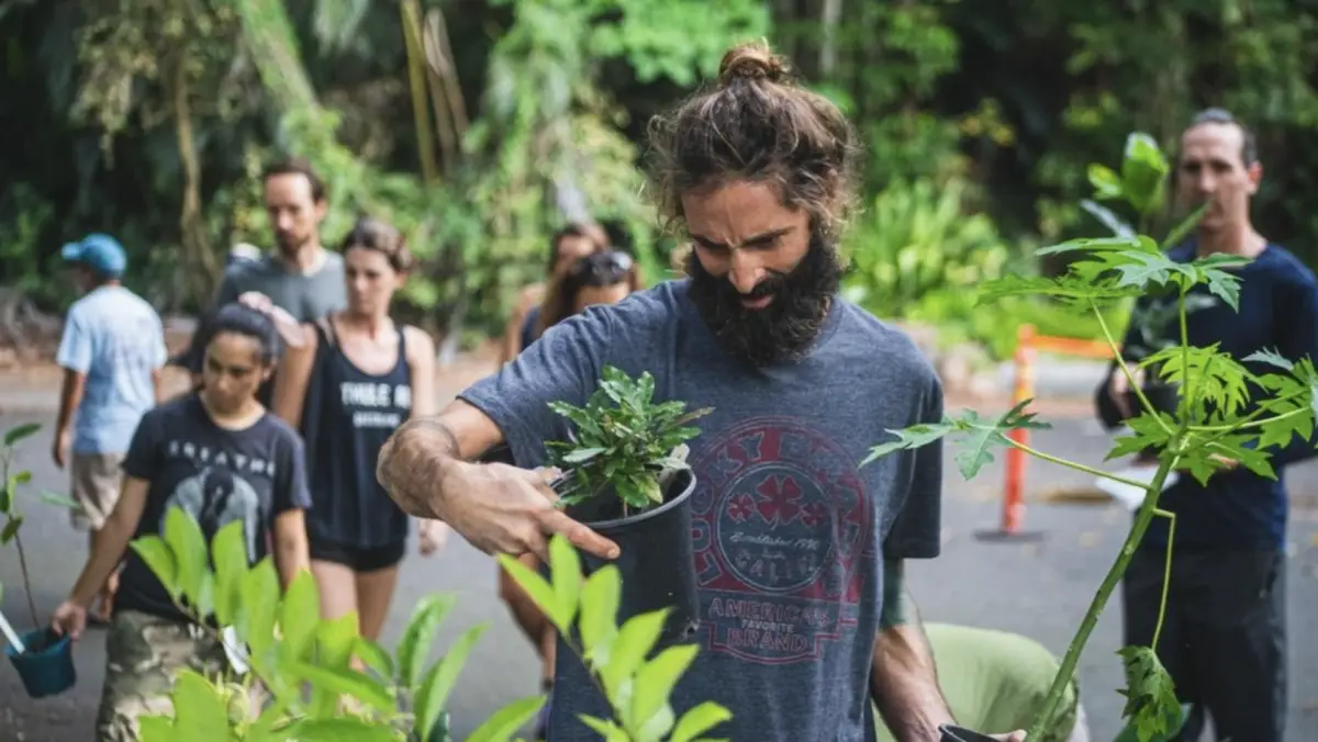 A man with a long beard holds a small potted plant while surrounded by greenery. He is accompanied by several people, some of whom are looking at plants. The scene appears to be set in an outdoor garden or nursery, possibly one of the charming North Shore annual events, with a lush, green background.