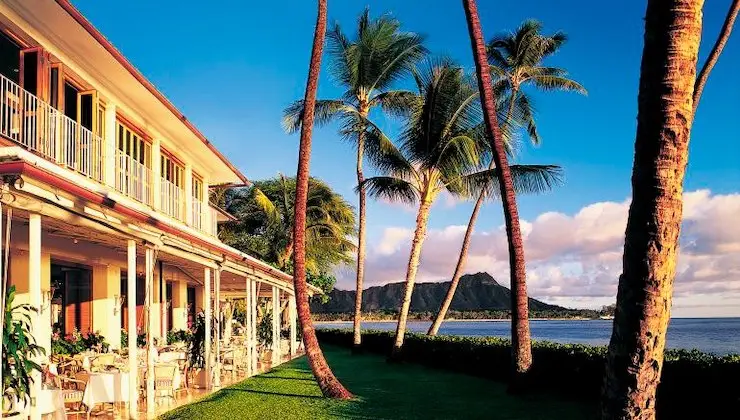 A picturesque scene featuring an elegant building with a balcony and outdoor dining area next to a vibrant green lawn, home to the best gluten-free dining in Waikiki. Tall palm trees sway gently, and in the background, there is a majestic mountain and serene ocean under a blue sky with soft clouds.