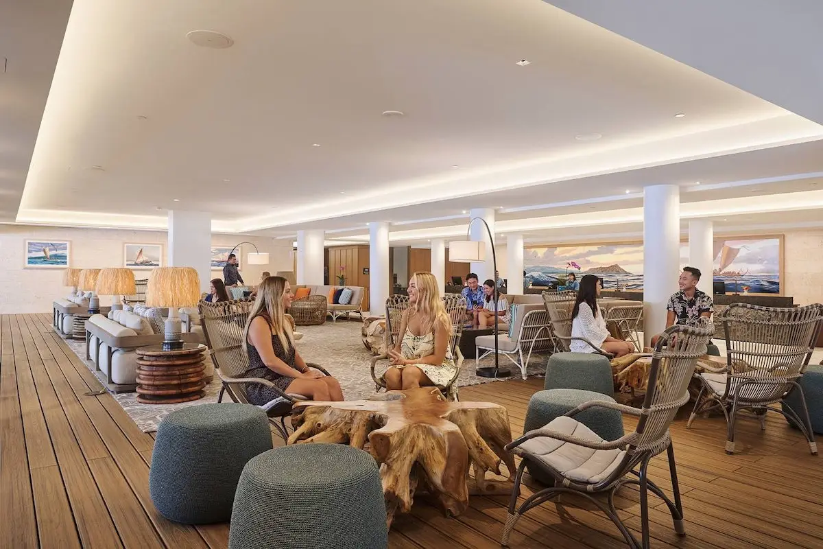 A spacious hotel lobby with a high ceiling and warm lighting, featuring several people sitting and talking on chairs and sofas. The decor includes wooden furniture, cushioned seating, and paintings on the walls, creating a cozy and inviting atmosphere reminiscent of Outrigger Hotels on Oahu.