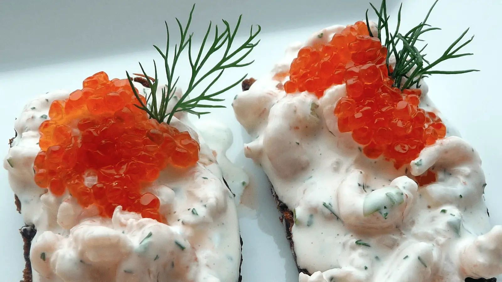 Two pieces of toast topped with a creamy white spread, garnished with vibrant orange roe (fish eggs) and sprigs of fresh dill. The dish is presented on a white plate, offering a visually appealing combination of colors and textures.