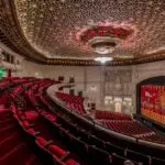 A luxurious theater interior showcasing an ornately decorated ceiling, red velvet seating, and elegant architectural details reminiscent of historic Bay Area theaters. The stage curtain features intricate designs, enhancing the venue's grandeur. The seating area curves towards the stage, providing an expansive view.