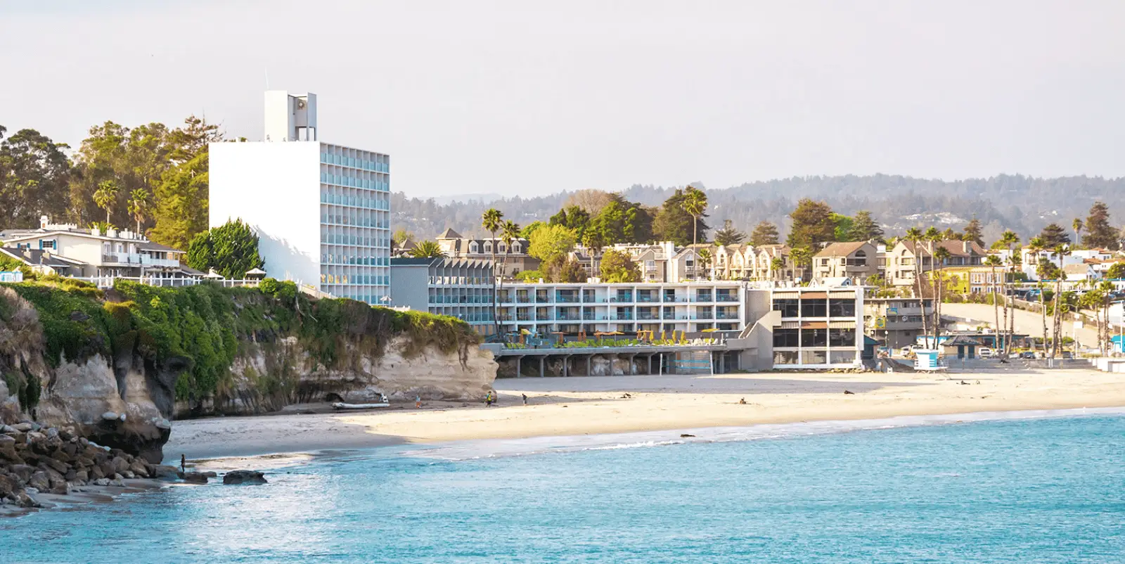 A scenic coastal view of a sandy beach bordered by turquoise waters showcases the benefits of life on the water. Cliffside buildings, including a tall white structure, are set against a backdrop of trees and residential areas. The beach and buildings are bathed in soft daylight, creating a serene atmosphere.