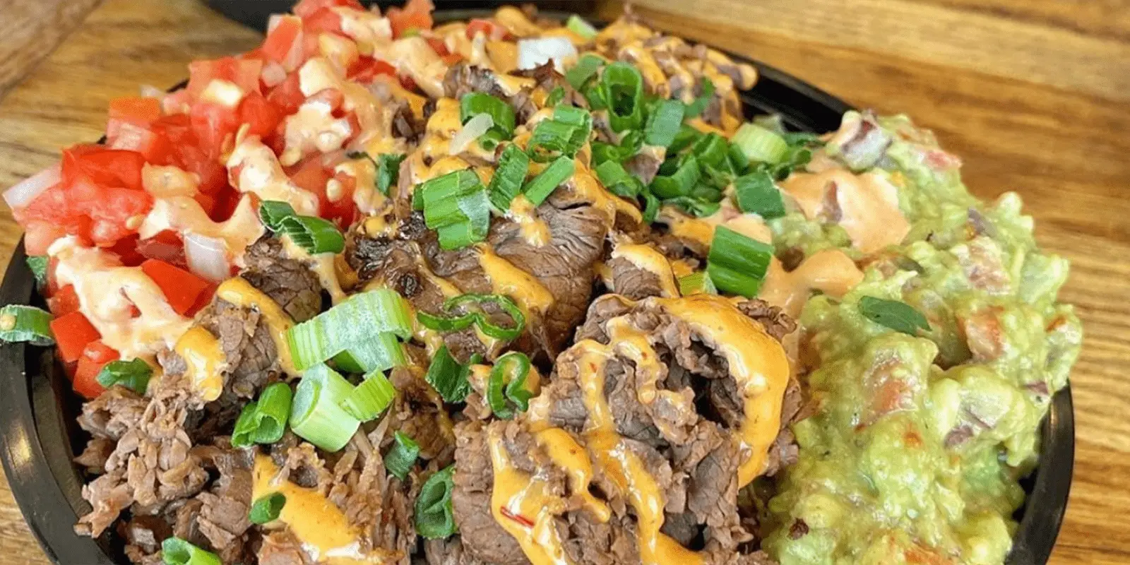 A close-up of a bowl filled with sliced grilled steak and topped with a drizzle of orange sauce and chopped green onions. The bowl, a highlight of the best gluten-free dining in Waikiki, also contains diced tomatoes, rice, and guacamole, all served on a wooden table.