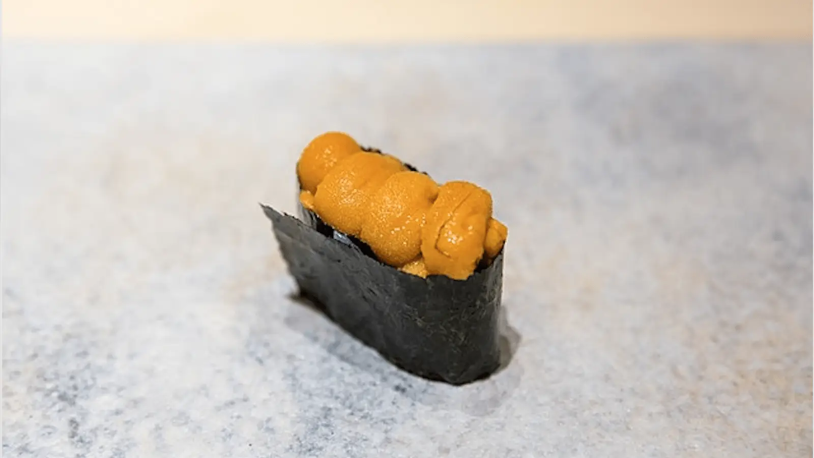 A piece of sushi featuring sea urchin (uni) placed over a small, oblong portion of white rice and wrapped partially in a strip of dark green seaweed (nori). The sushi is resting on a light grey surface.
