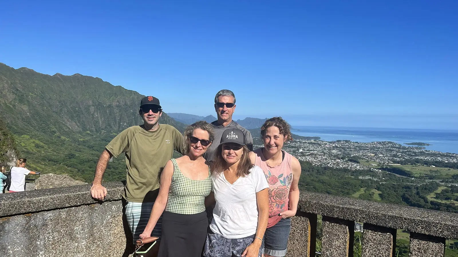 Five people are posing for a photo at a scenic overlook with lush green mountains and a coastal town in the background under a clear blue sky. They are standing behind a stone railing, looking happy and relaxed, capturing memories of their adventure. Why Waikiki? It's simply breathtaking!