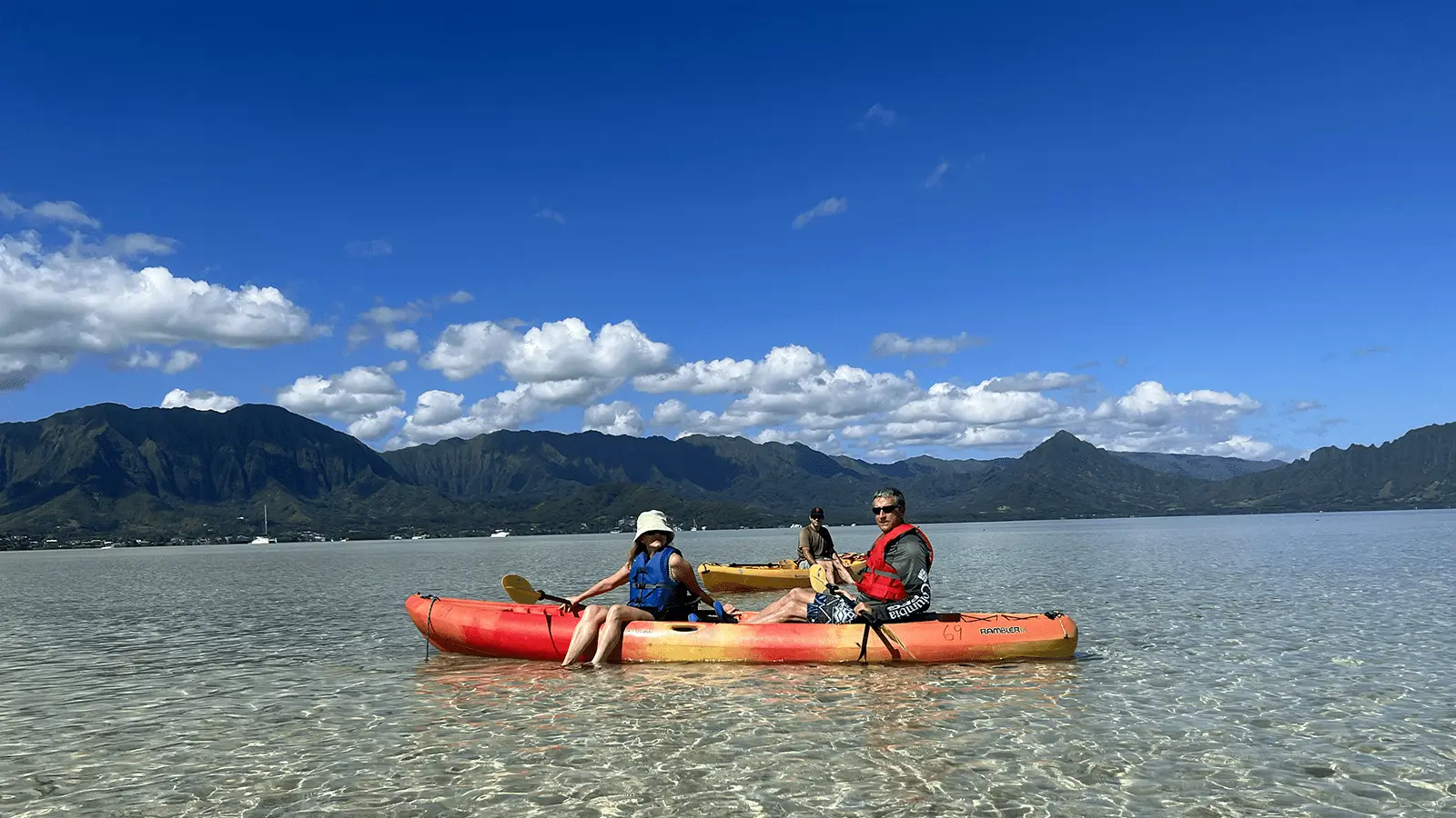 Two people and a dog are sitting in a bright orange kayak on clear, shallow water. The individuals are wearing life jackets, and the person on the left is holding a paddle. Behind them, there are picturesque mountains under a blue sky with scattered clouds—a perfect scene that makes you wonder: Why Waikiki?