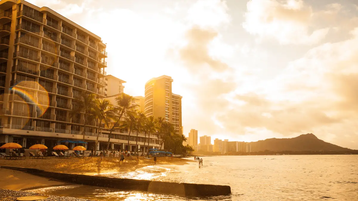 A scenic, golden sunset view of Waikiki beachfront lined with high-rise buildings. People are seen enjoying the beach, with the shoreline illuminated by the setting sun. The sky is partly cloudy, casting a warm glow over the landscape, with mountains in the background. Why Waikiki?